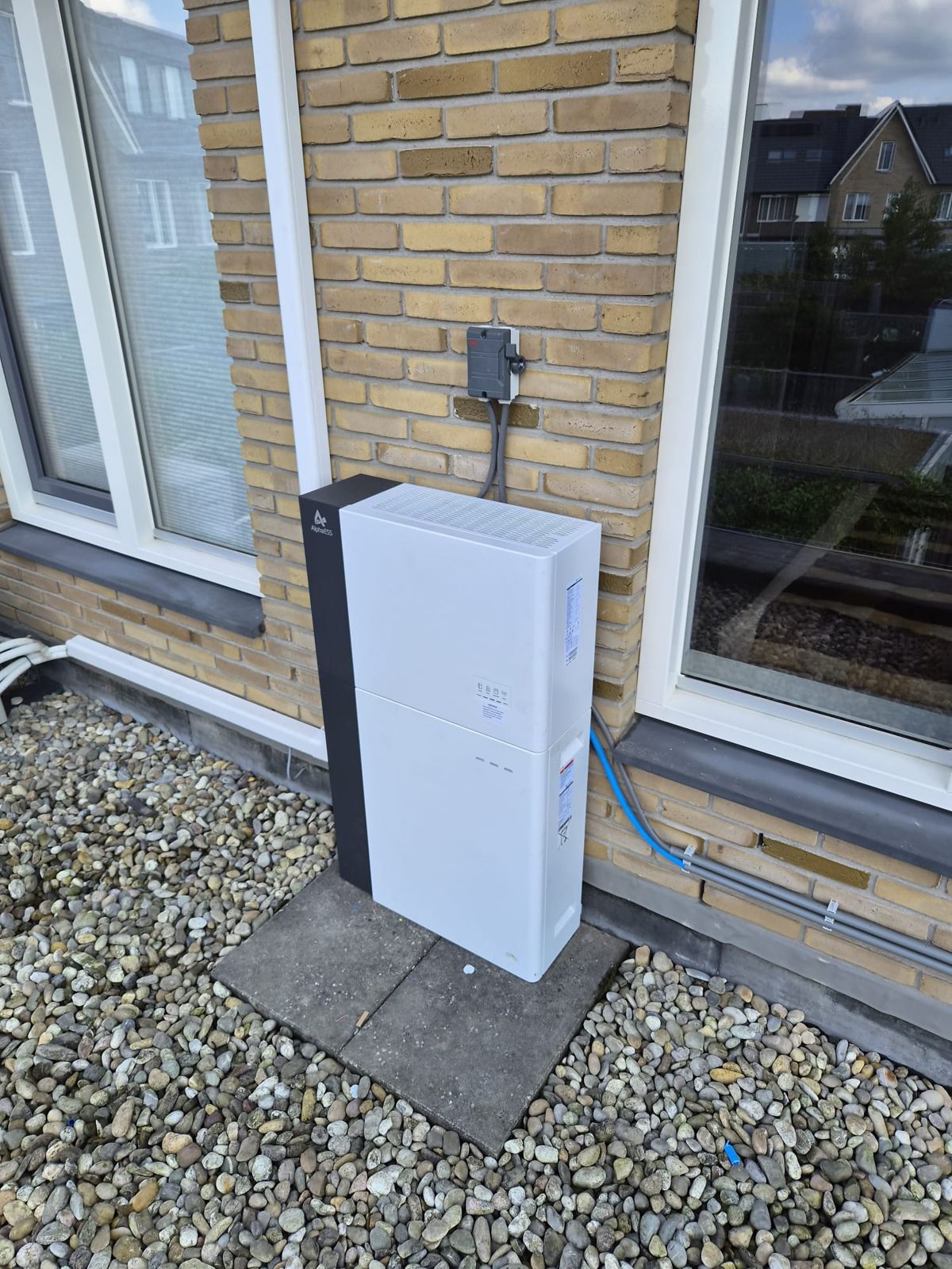 AlphaESS-thuisbatterij buiten tegen een bakstenen gevel, op een tegel in het grind